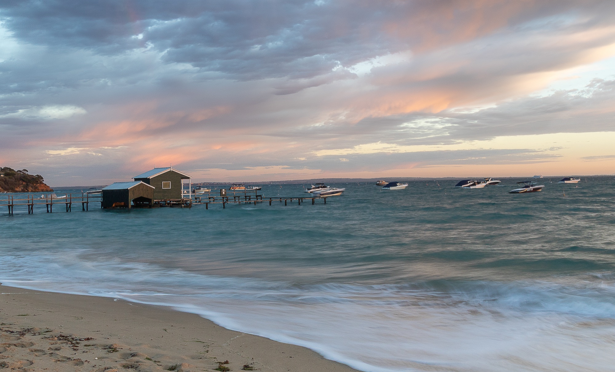 Boatshed on a pier at sunset, anchored boats in the bay.